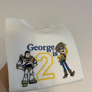 Może przedstawiać: Biała bluza z haftowanymi postaciami z Toy Story, Buzzem Astralem i Chudym. Tekst "George is 2" jest wyhaftowany na niebiesko i ż&oacute;łto. Bluza jest trzymana przez rękę.