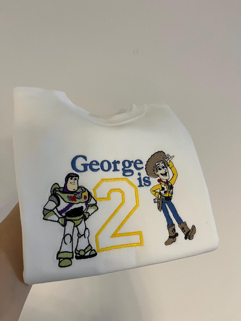 Może przedstawiać: Biała bluza z haftowanymi postaciami z Toy Story, Buzzem Astralem i Chudym. Tekst "George is 2" jest wyhaftowany na niebiesko i ż&oacute;łto. Bluza jest trzymana przez rękę.