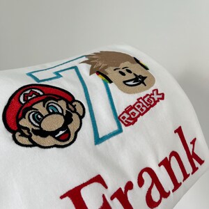 May include: White fabric with embroidered designs. Features a cartoon image of Mario in red and tan, a Roblox character, the number 7 in teal, and the name "Frank" in red.