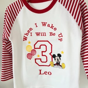 Könnte beinhalten: Weißes und rotes Raglan-Shirt mit rot gestreiften Ärmeln. Das Shirt zeigt den Text "When I Wake I Will Be Up 3 Leo" in roter Stickerei, zusammen mit einer Mickey Mouse-Grafik und Luftballons.