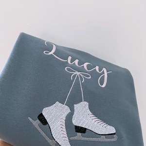 以下が含まれることがあります： 薄い青色のスウェットシャツに「Lucy」という名前が刺繍されています。スウェットシャツには、白いリボンからぶら下がった、黒いブレードが付いた白いスケート靴が描かれています。