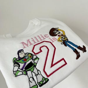 Może przedstawiać: Biała bluza z haftowanymi postaciami Buzza Astrala i Chudego z Toy Story. Tekst "Millie is 2" jest wyhaftowany na r&oacute;żowo i czerwono. Odzież dziecięca, idealna na drugie urodziny.