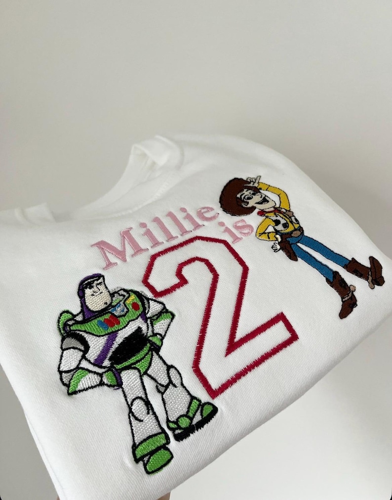Może przedstawiać: Biała bluza z haftowanymi postaciami Buzza Astrala i Chudego z Toy Story. Tekst "Millie is 2" jest wyhaftowany na r&oacute;żowo i czerwono. Odzież dziecięca, idealna na drugie urodziny.