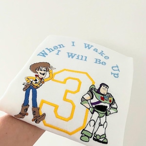 Puede incluir: Camiseta blanca con imágenes bordadas de Woody y Buzz Lightyear de Toy Story. La camiseta también tiene el número 3 y el texto "When I Wake I Will Be Up".