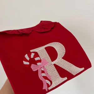 May include: Red sweatshirt with a white embroidered letter "R" and a candy cane with a pink bow. The sweatshirt has a scalloped collar.