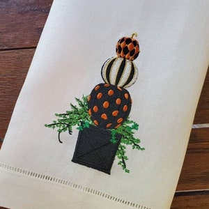 May include: A white linen kitchen towel with an embroidered design of a stack of three pumpkins in a black pot. The pumpkins are orange, black, and white with polka dots. The pot is surrounded by green leaves.