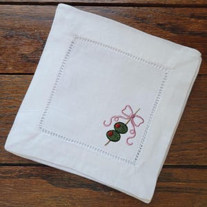 May include: White linen cocktail napkin with a decorative embroidered design. The design features two green olives with red centers on a wooden skewer, accented by a pink bow. The napkin has a decorative stitched border.