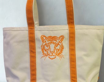 May include: Cream-colored canvas tote bag with orange accents. Features an embroidered orange tiger face on the front. The bag has orange handles and a matching orange base. A stylish and practical accessory for carrying essentials.