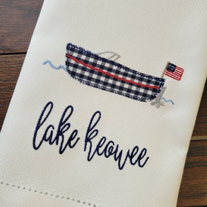 May include: White kitchen towel with blue and red embroidered boat and the text "Lake Keowee".