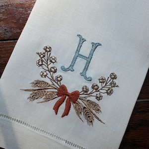 May include: A white linen tea towel with an embroidered wreath of wheat and berries, a red bow, and the letter "H" in blue thread.