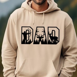 May include: A tan hoodie with a front pocket and drawstring hood. The word "DAD" is printed across the chest in a black, stylized font, with a silhouette of an archer, deer, and deer in the letters.