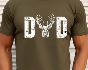 May include: Olive green t-shirt with the word "DAD" in distressed white letters, featuring a deer head graphic in the center. The shirt is a short-sleeved crew neck.