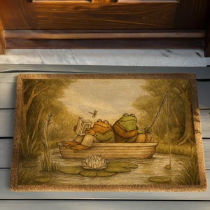 May include: A rectangular doormat featuring a whimsical illustration of two frogs in a boat. One frog reads a book, while the other fishes. The scene is set in a pond with lily pads and trees, with a natural fiber border.