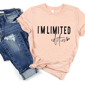 May include: A peach-colored t-shirt with the text "I'M LIMITED edition" in black. The shirt is styled with a knot at the bottom and paired with blue denim jeans with distressed details.