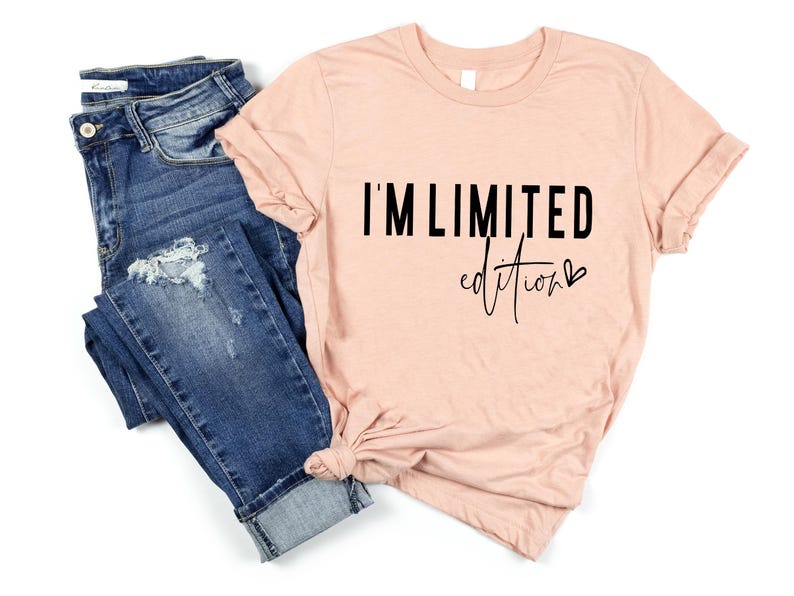 May include: A peach-colored t-shirt with the text "I'M LIMITED edition" in black. The shirt is styled with a knot at the bottom and paired with blue denim jeans with distressed details.