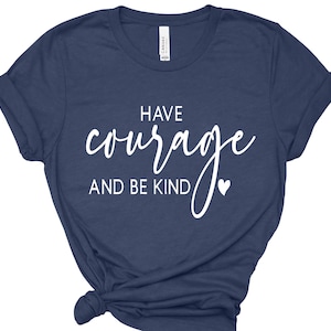 May include: Navy blue t-shirt with the words "HAVE courage AND BE KIND" in white script. A small white heart is next to the word "KIND". The shirt is folded at the bottom.