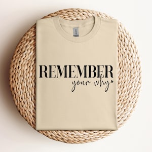 May include: A beige t-shirt with the words "REMEMBER your why" printed in black. The shirt is folded and resting on a round, woven wicker placemat. The text is in a bold, sans-serif font with a cursive "your why".