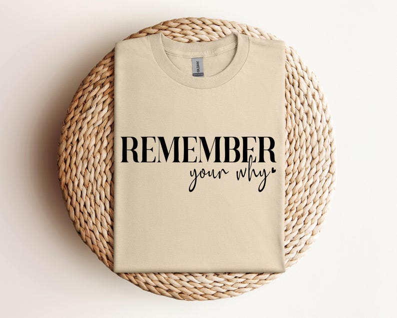 May include: A beige t-shirt with the words "REMEMBER your why" printed in black. The shirt is folded and resting on a round, woven wicker placemat. The text is in a bold, sans-serif font with a cursive "your why".
