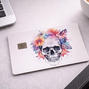 May include: A white credit card with a skull design decorated with colourful flowers. The skull is central, with orange, pink, and blue floral elements. A small chip is visible on the card.