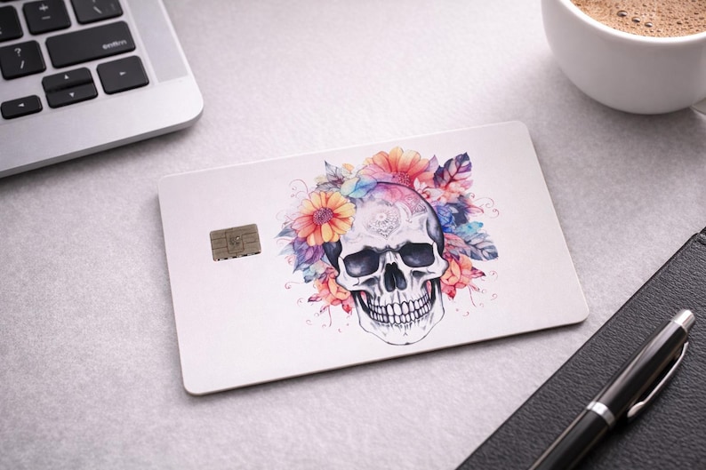May include: A white credit card with a skull design decorated with colourful flowers. The skull is central, with orange, pink, and blue floral elements. A small chip is visible on the card.