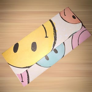 Puede incluir: Un sobre de papel plegado con un patrón repetido de caras sonrientes en amarillo, rosa, azul y melocotón. Los contornos y detalles negros de las caras contrastan con los colores pastel. El sobre está sobre una superficie de madera marrón claro.