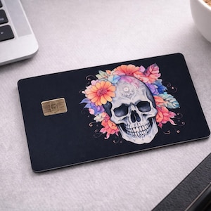 May include: Black card featuring a skull design with orange, pink, and purple floral accents. The skull is white. A gold-coloured chip is visible. The card is on a light grey surface.