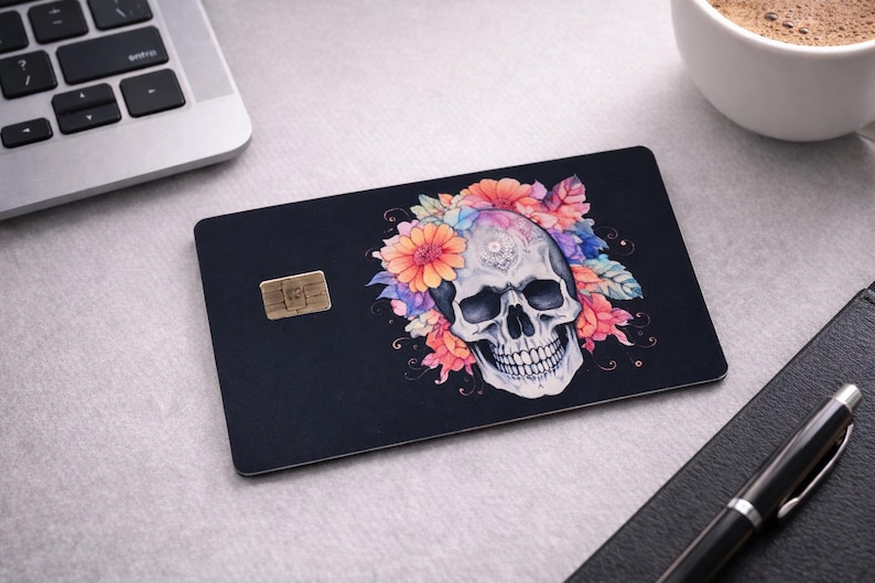 May include: Black card featuring a skull design with orange, pink, and purple floral accents. The skull is white. A gold-coloured chip is visible. The card is on a light grey surface.