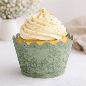 May include: A single cupcake with white frosting and gold sprinkles sits in a decorative green paper cup. The cup features a pastoral scene with trees and figures. The cupcake is on a white surface.