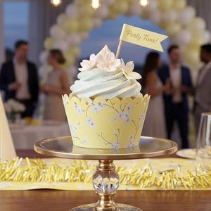 May include: A decorated cupcake with light blue frosting and white flower accents sits on a gold cake stand. The yellow cupcake liner has a floral design. A small flag that says "Party Time!" is on top. Party decorations are in the background.