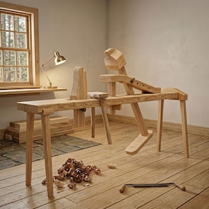 Può includere: Un cavallino di legno, uno strumento per la lavorazione del legno, è il punto focale di questa immagine. L'utensile in legno chiaro si trova in un'officina, con trucioli di legno sul pavimento. Una piccola lampada e una finestra sono sullo sfondo.