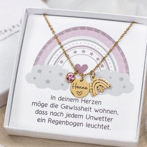 May include: A gold necklace with a rainbow charm, a small pink crystal, and a round charm with the name "Hanna" and a heart. The necklace is in a white box with a pink and gray rainbow design and a message that reads "In your heart may the certainty live that after every storm a rainbow shines."
