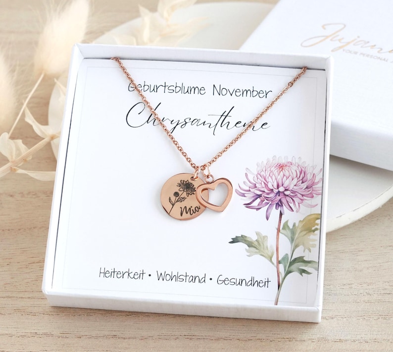 May include: A rose gold necklace with a chrysanthemum charm and a heart charm. The necklace comes in a white box with a card that says "Geburtsblume November Chrysantheme" and "Heiterkeit Wohlstand Gesundheit".