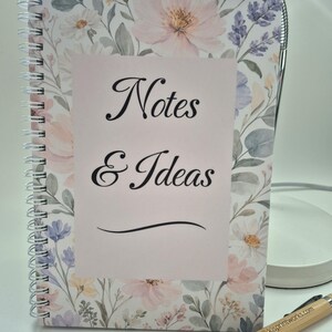 May include: A floral notebook with a spiral binding, displaying the words "Notes & Ideas" in black script. The design includes pink and white flowers. A wooden pen with silver details is placed nearby.