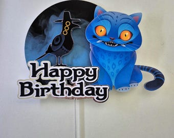 May include: A blue cat cake topper with orange eyes and a toothy grin. A black raven with a hat is on a dark blue background. The words "Happy Birthday" are in black and white.