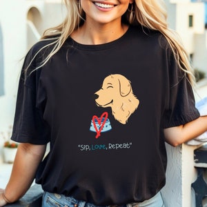 Op de afbeelding: Zwart T-shirt met een afbeelding van een golden retriever die naar een rood hart boven een blauwe hondenkom kijkt. De tekst "Sip, Love, Repeat" staat onder de afbeelding.