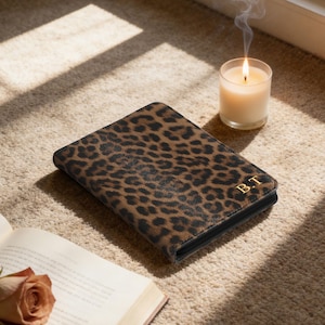 Op de afbeelding: Een notitieboek met luipaardprint en gouden initialen "B.T" ligt op een beige tapijt. Een brandende kaars in een glazen pot en een open boek met een roos zijn ook zichtbaar. De kaft van het notitieboek heeft een bruin en zwart vlekkenpatroon.