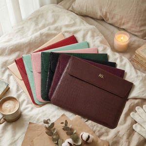 Puede incluir: Una colección de fundas para portátiles texturizadas y coloridas en varios tonos, incluyendo rojo, verde y marrón. La funda marrón está grabada con las iniciales "JG". Los artículos están dispuestos sobre una cama con un libro, café y una vela encendida.