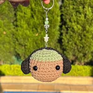 Könnte beinhalten: Ein handgefertigter Häkel-Schlüsselanhänger mit einem runden, hellbraunen Kopf mit schwarzen Ohren und einem hellgrünen Oberteil. Der Schlüsselanhänger hat schwarze Knopfaugen und einen silbernen Ring mit einem Perlenanhänger. Der Anhänger enthält grüne Perlen und einen Stern.