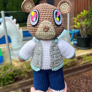 Puede incluir: Una muñeca de oso de ganchillo hecha a mano con cabeza marrón, orejas color canela y ojos grandes con detalles en rosa, amarillo y azul. El oso lleva una camisa blanca, un chaleco gris y pantalones azules. La muñeca se sostiene en una mano.
