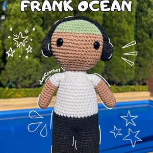 Pode incluir: Padrão de crochê para uma boneca amigurumi de Frank Ocean. A boneca está vestindo uma camisa branca, calças pretas e um gorro verde. A boneca está segurando um microfone em sua mão direita.