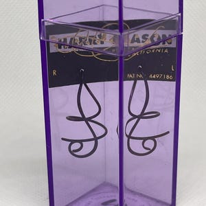 May include: A pair of dark, thin, spiral-shaped wire earrings displayed inside a tall, transparent purple plastic hexagonal case. The black backing card inside the case features the text "HARRY MASON CALIFORNIA" and "PAT NO 4497186".