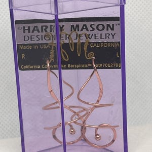 May include: A pair of copper-colored metal earspirals by Harry Mason Designer Jewelry, labeled California Convertible Earspirals, inside a transparent purple plastic rectangular case. The jewelry features a continuous spiral wire design.