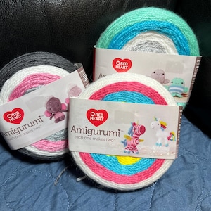 May include: Three skeins of Red Heart Amigurumi yarn in various color combinations. One skein features black, gray, pink, and white. Another has white, pink, blue, and yellow. The third has shades of blue, white, and gray. Each skein has a label with the text "Amigurumi".