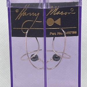 May include: A pair of silver-toned earrings featuring a curved wire design and a single dark gray spherical bead, displayed on a black card labeled Harry Mason with Pat. No. 1497186, enclosed within a transparent purple plastic protective case.