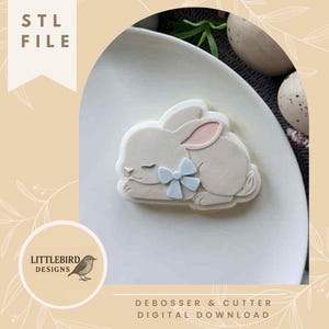 May include: A light-colored bunny cookie with a blue bow, resting on a white plate. The cookie has pink inner ears and closed eyes. The image includes the text "STL FILE" and "DEBOSSER & CUTTER DIGITAL DOWNLOAD".