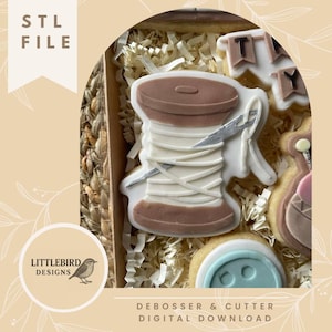 Può includere: Una scatola di biscotti decorati, tra cui una spoletta di filo, un bottone e biscotti a forma di banner. I biscotti sono bianchi, marroni e blu, con dettagli intricati. La scatola contiene imbottitura di carta e il testo "STL FILE" e "DEBOSSER & CUTTER DIGITAL DOWNLOAD".