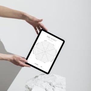 Może przedstawiać: Czarny tablet z białym tłem, na kt&oacute;rym widoczne jest koło audytu życia z kategoriami takimi jak zdrowie, finanse i rozw&oacute;j osobisty. Tablet jest trzymany przez ręce i spoczywa na białym kamieniu.