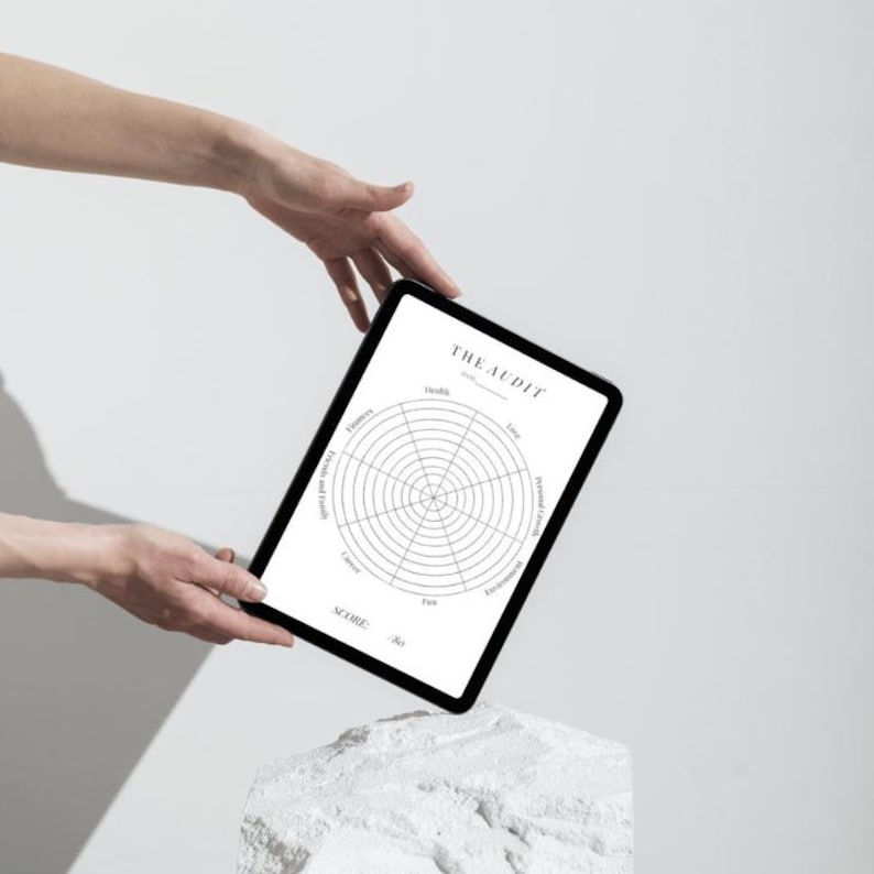 Może przedstawiać: Czarny tablet z białym tłem, na kt&oacute;rym widoczne jest koło audytu życia z kategoriami takimi jak zdrowie, finanse i rozw&oacute;j osobisty. Tablet jest trzymany przez ręce i spoczywa na białym kamieniu.