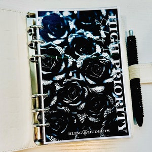 May include: White leather binder with a floral insert featuring black roses and the text "HIGH PRIORITY" and "BLING & BUDGETS." A black pen with a white band rests beside the binder on a white surface.