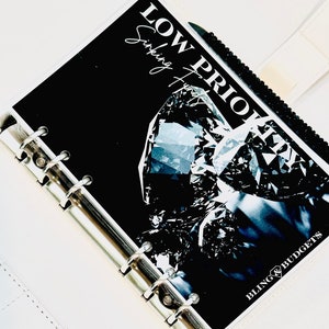 May include: A white leather planner with a black and white insert that says "LOW PRIORITIES" and "Surfing the" with a diamond image. The planner has a silver ring binder.